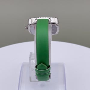 May include: A silver wristwatch with a square face and a green leather strap. The watch is displayed on a clear stand, highlighting the strap's colour and the watch's design. The strap is approximately 18 cm long.