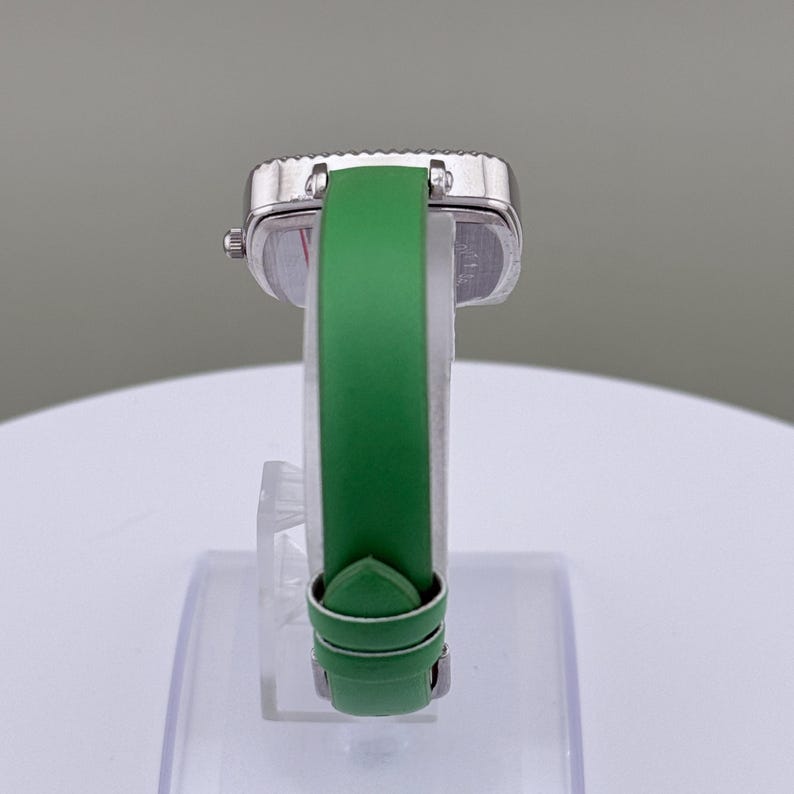 May include: A silver wristwatch with a square face and a green leather strap. The watch is displayed on a clear stand, highlighting the strap's colour and the watch's design. The strap is approximately 18 cm long.