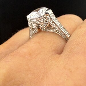 May include: A silver ring with a large, clear, round-cut gemstone as the centerpiece. The band is encrusted with smaller, clear gemstones. The ring is set against a dark background.
