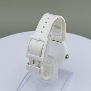 Puede incluir: Un reloj de pulsera blanco con correa y hebilla a juego. El reloj se exhibe sobre un soporte de pl&aacute;stico transparente, mostrando los detalles de la correa y la esfera del reloj. El reloj tiene un dise&ntilde;o simple y minimalista.