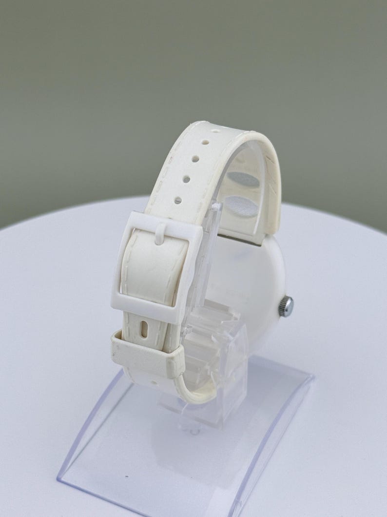 Puede incluir: Un reloj de pulsera blanco con correa y hebilla a juego. El reloj se exhibe sobre un soporte de pl&aacute;stico transparente, mostrando los detalles de la correa y la esfera del reloj. El reloj tiene un dise&ntilde;o simple y minimalista.