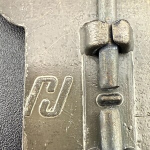 May include: Close-up of a weathered metal latch with a sliding bolt mechanism. The latch features a stylized "H" logo and is likely part of a door or cabinet hardware. The metal has a worn, industrial appearance.