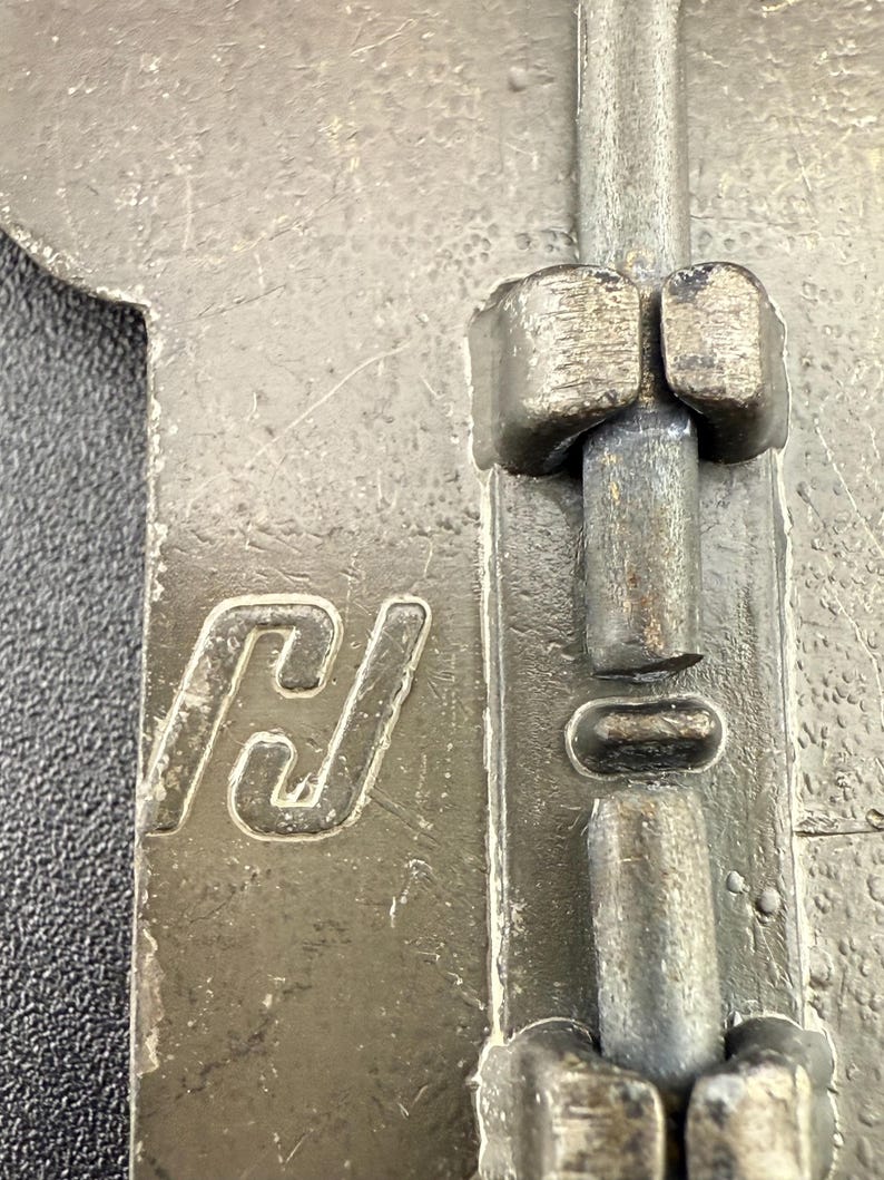 May include: Close-up of a weathered metal latch with a sliding bolt mechanism. The latch features a stylized "H" logo and is likely part of a door or cabinet hardware. The metal has a worn, industrial appearance.