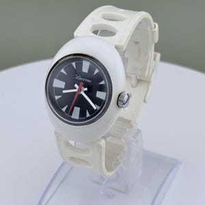 Puede incluir: Reloj Lucerna blanco con esfera negra y marcadores blancos. El reloj tiene una correa blanca con detalles recortados. La esfera del reloj tiene la palabra "Lucerna" impresa. El reloj se exhibe sobre un soporte transparente.
