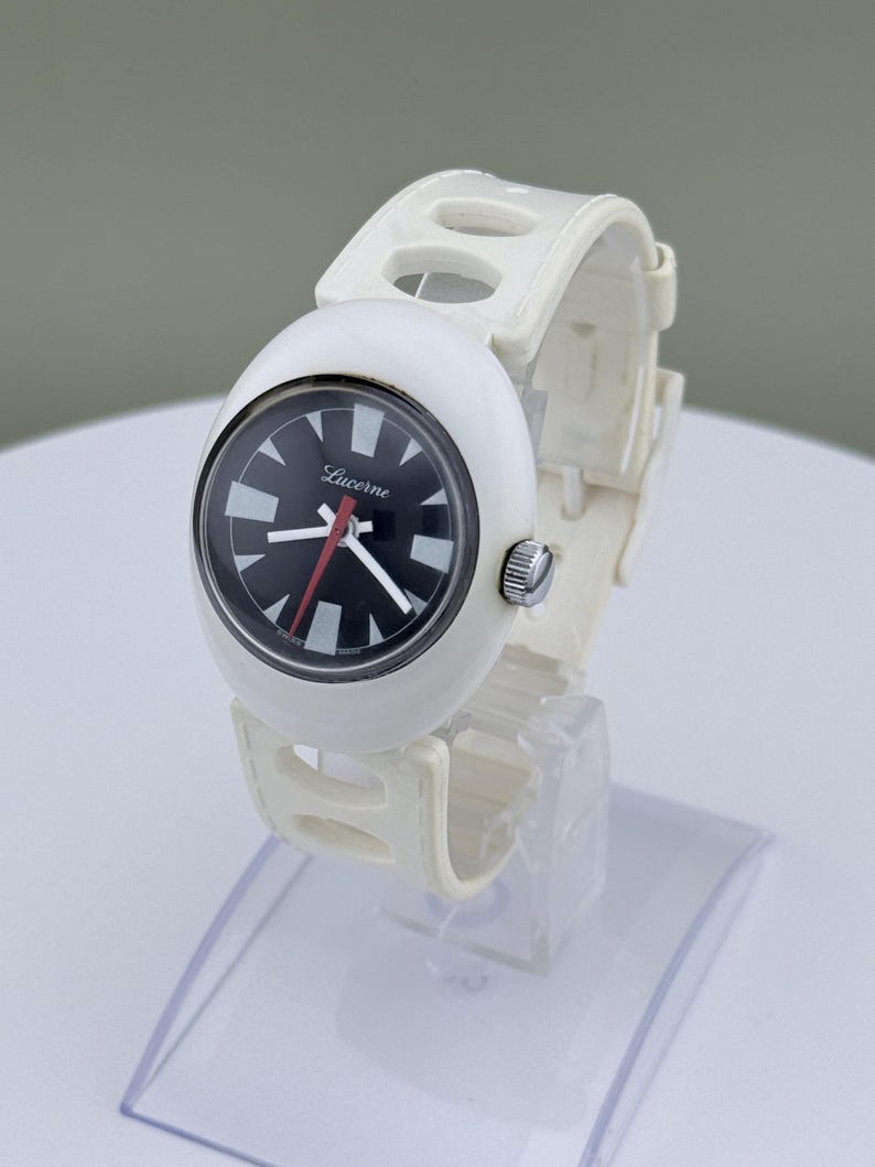 Puede incluir: Reloj Lucerna blanco con esfera negra y marcadores blancos. El reloj tiene una correa blanca con detalles recortados. La esfera del reloj tiene la palabra "Lucerna" impresa. El reloj se exhibe sobre un soporte transparente.