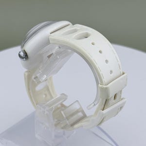 Puede incluir: Un reloj de pulsera blanco con una correa blanca. La esfera del reloj tiene un dise&ntilde;o a rayas grises y blancas claras. El reloj se exhibe en un soporte de pl&aacute;stico transparente.