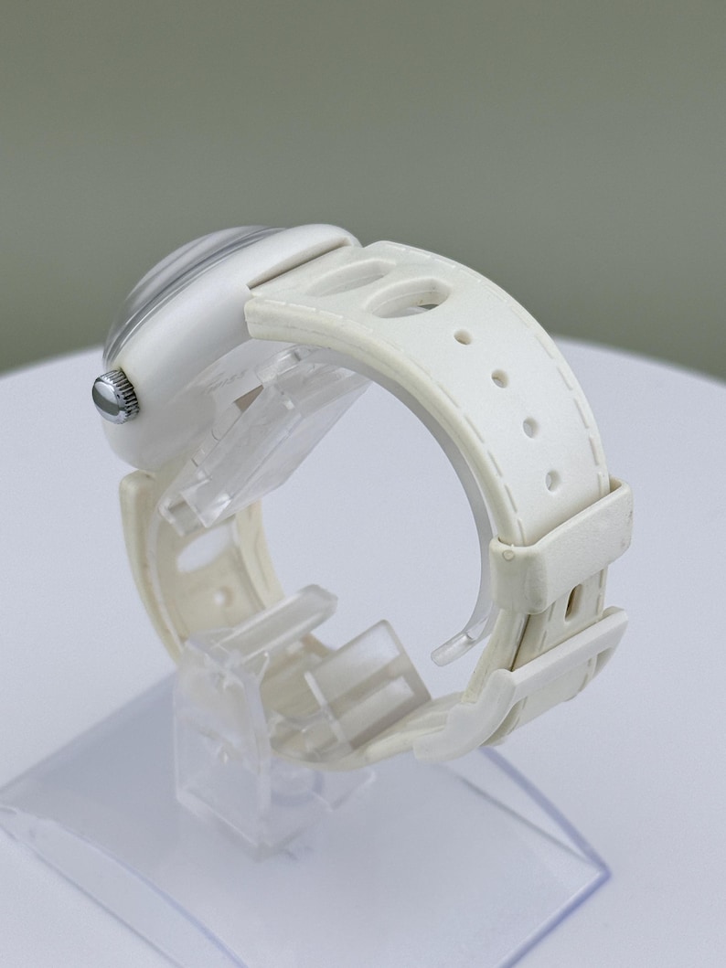 Puede incluir: Un reloj de pulsera blanco con una correa blanca. La esfera del reloj tiene un dise&ntilde;o a rayas grises y blancas claras. El reloj se exhibe en un soporte de pl&aacute;stico transparente.