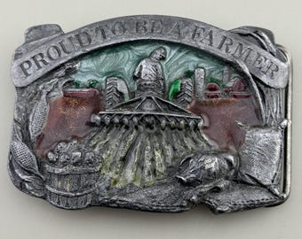 May include: A silver-toned metal belt buckle with the words "PROUD TO BE A FARMER" arched across the top. The buckle features a detailed scene of a farmer, tractor, and farm buildings, with accents of red, green, and brown.