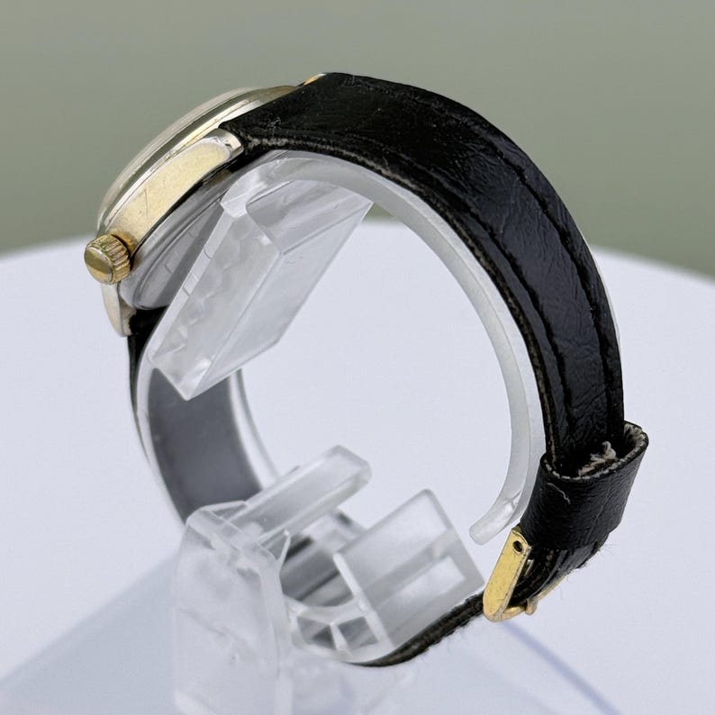 K&ouml;nnte beinhalten: Eine Vintage-Armbanduhr mit einem goldfarbenen Geh&auml;use und einem schwarzen Lederarmband. Die Uhr hat ein klassisches Design mit rundem Zifferblatt und goldfarbener Krone. Das Armband wird mit einer goldfarbenen Schnalle befestigt.