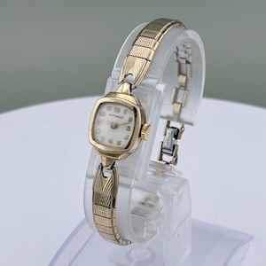 May include: A vintage gold-toned Wittnauer wristwatch with a rectangular face and a gold-toned, expandable band. The watch face has a cream-colored background with black numerals and hands. The watch is displayed on a clear stand.