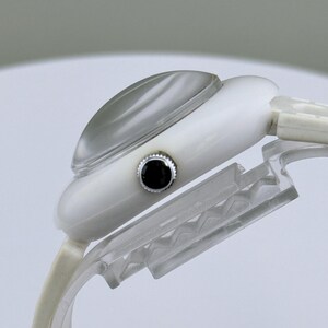 Puede incluir: Un reloj de pulsera blanco con un cristal curvo transparente y un bot&oacute;n negro. El reloj se muestra sobre un soporte de pl&aacute;stico transparente, mostrando su perfil lateral. La correa del reloj es blanca.