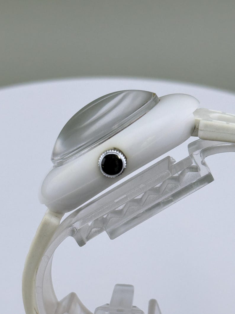 Puede incluir: Un reloj de pulsera blanco con un cristal curvo transparente y un bot&oacute;n negro. El reloj se muestra sobre un soporte de pl&aacute;stico transparente, mostrando su perfil lateral. La correa del reloj es blanca.