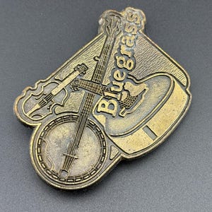 May include: A bronze-colored metal belt buckle featuring a banjo, guitar, and violin. The word "Bluegrass" is embossed across the top. The buckle has a detailed design and is set against a dark background.