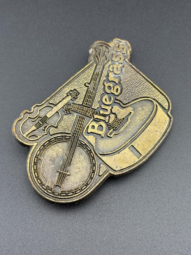 May include: A bronze-colored metal belt buckle featuring a banjo, guitar, and violin. The word "Bluegrass" is embossed across the top. The buckle has a detailed design and is set against a dark background.