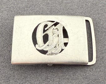 May include: Silver-toned belt buckle with an engraved design. The buckle features a rectangular shape with a loop on one end and a decorative oval cutout in the center. The cutout reveals an ornate monogram of the letters "GM".
