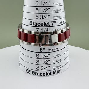 May include: A red and silver bracelet is displayed on a white and black EZ Bracelet Mini sizing tool. The bracelet is approximately 7 inches (17.8 cm) in length. The bracelet has a rectangular link design.