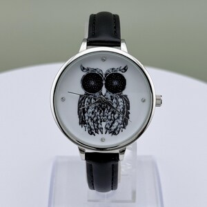 May include: A silver-toned wristwatch with a black leather band. The watch face features a black and white owl illustration. The watch has a round face with small crystal accents and a silver crown.