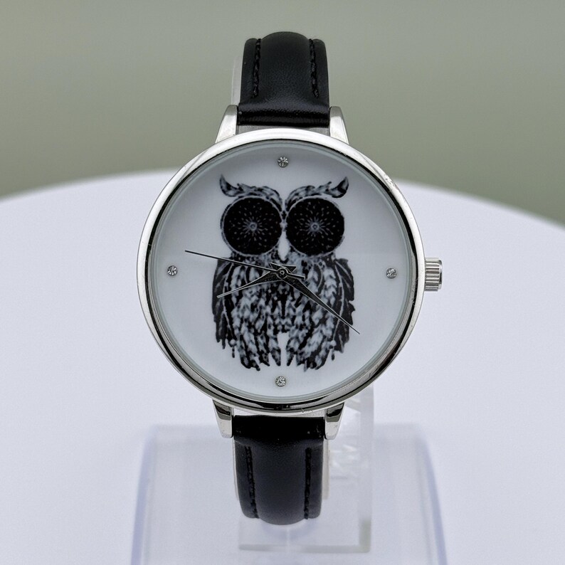 May include: A silver-toned wristwatch with a black leather band. The watch face features a black and white owl illustration. The watch has a round face with small crystal accents and a silver crown.