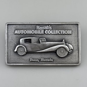 May include: A rectangular, silver-colored belt buckle featuring a vintage car design. The buckle is embossed with the text "Harrah's AUTOMOBILE COLLECTION" and "Reno, Nevada". The car has detailed wheels and body.
