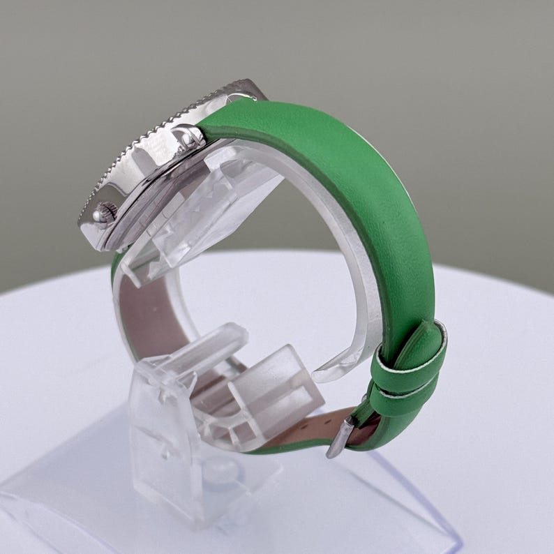 May include: A silver wristwatch with a green leather strap. The watch features a textured bezel. The watch is displayed on a clear stand, highlighting the green strap and the watch's side profile.