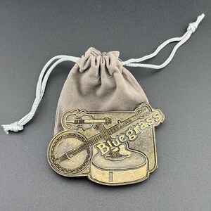 May include: A bronze-colored metal banjo and fiddle pin with the word "Bluegrass" on it, resting on a small, tan drawstring bag. The pin features detailed instrument designs, perfect for music lovers.