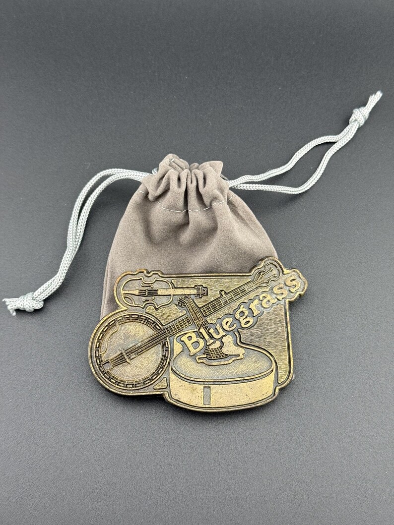 May include: A bronze-colored metal banjo and fiddle pin with the word "Bluegrass" on it, resting on a small, tan drawstring bag. The pin features detailed instrument designs, perfect for music lovers.