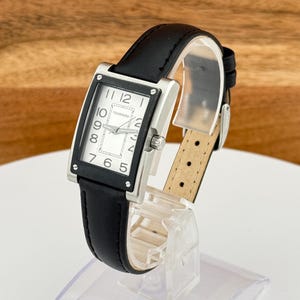May include: A rectangular Tourneau watch with a black leather band. The watch face is white with black numerals and hands. The watch has a silver-colored bezel and a black leather strap.