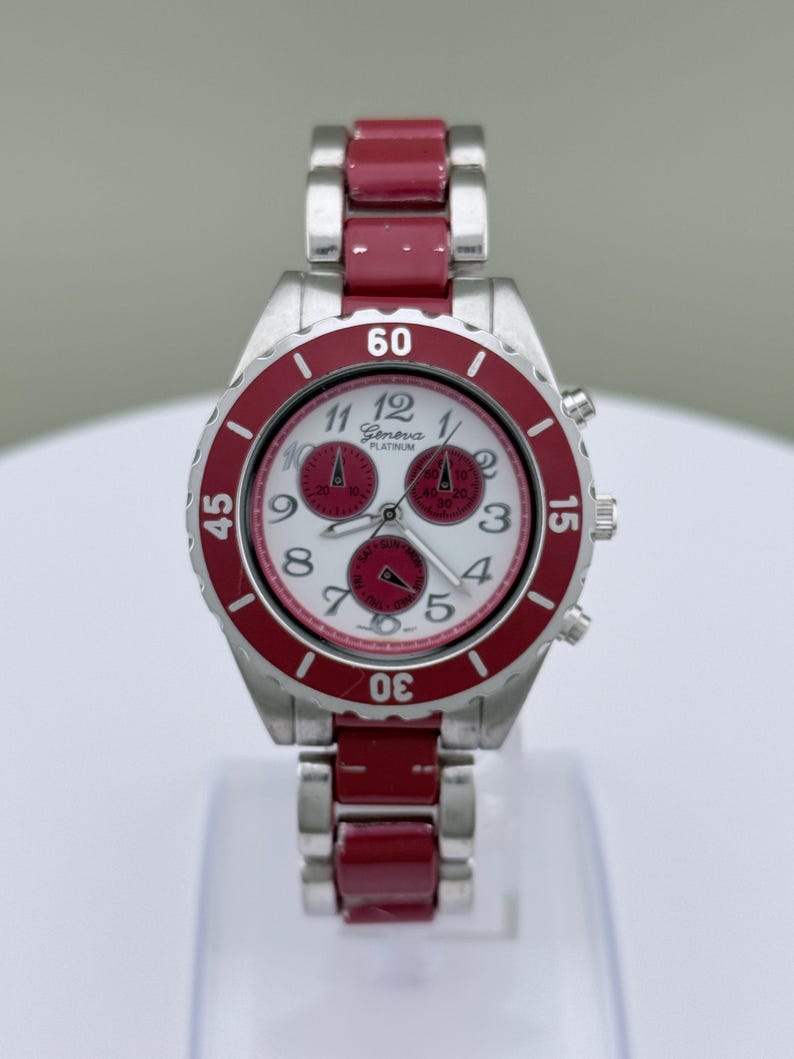 May include: A Geneva Platinum wristwatch with a silver-tone case and a red bezel and band. The watch face is white with red sub-dials and black numerals. The watch has a chronograph design.