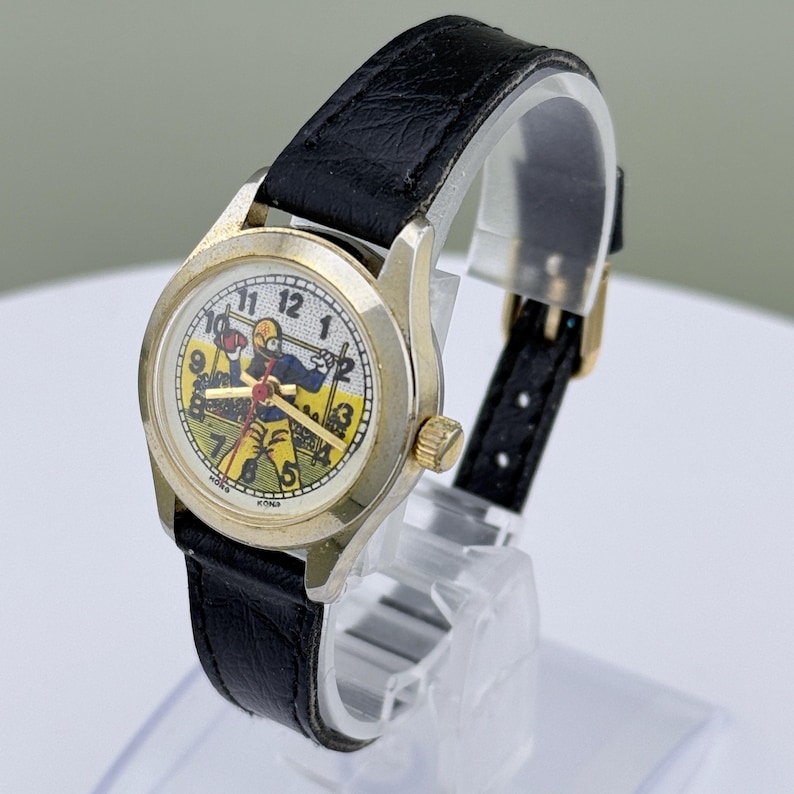 K&ouml;nnte beinhalten: Vintage Armbanduhr mit goldfarbenem Geh&auml;use und schwarzem Lederarmband. Das Zifferblatt der Uhr zeigt eine farbenfrohe Illustration eines Footballspielers. Die Uhr hat eine goldene Krone und ein schwarzes Lederarmband.