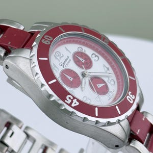 May include: A Geneva Platinum wristwatch with a white face and red accents. The watch features a silver-tone metal band and a red bezel with white numerals. The watch face has three sub-dials and silver hands.