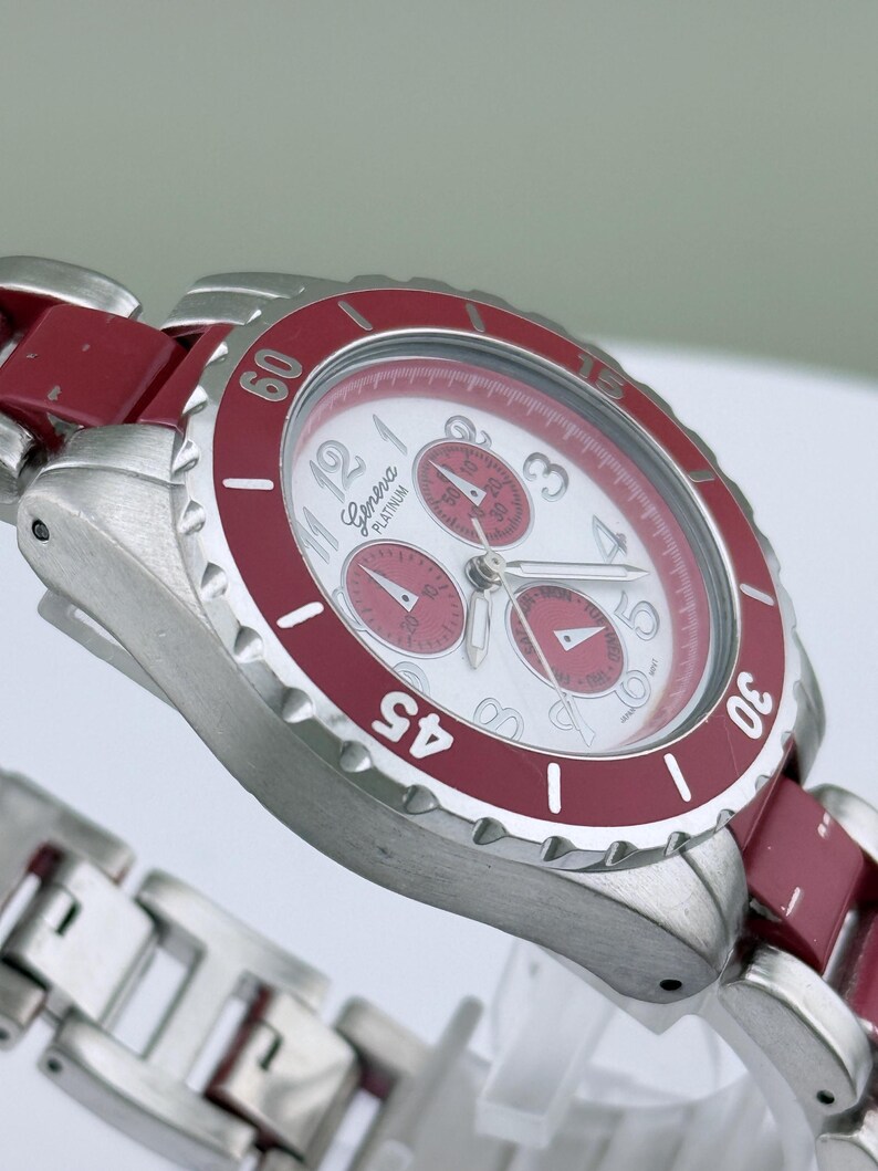 May include: A Geneva Platinum wristwatch with a white face and red accents. The watch features a silver-tone metal band and a red bezel with white numerals. The watch face has three sub-dials and silver hands.