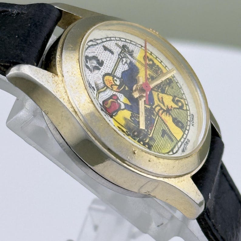 K&ouml;nnte beinhalten: Eine Vintage-Armbanduhr mit einem goldfarbenen Geh&auml;use und einem schwarzen Lederarmband. Das Zifferblatt der Uhr zeigt ein farbenfrohes Cartoon-Charakter-Design mit roten Zeigern. Die Uhr ist abgewinkelt und zeigt die Seite und das Zifferblatt.