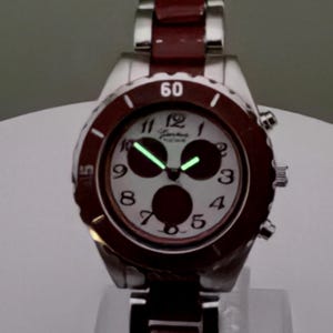 May include: A silver wristwatch with a burgundy bezel and band. The watch face is white with black numerals and three dark circles. The hands and numerals glow green. The watch is displayed on a clear stand.