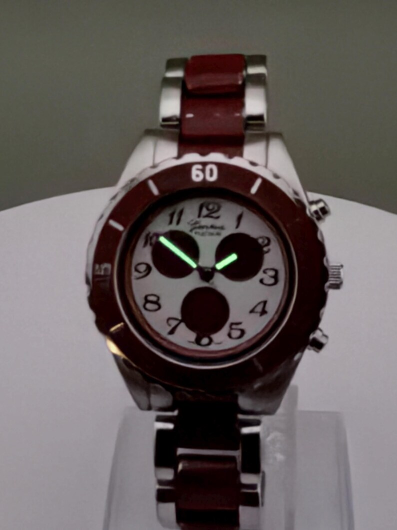 May include: A silver wristwatch with a burgundy bezel and band. The watch face is white with black numerals and three dark circles. The hands and numerals glow green. The watch is displayed on a clear stand.