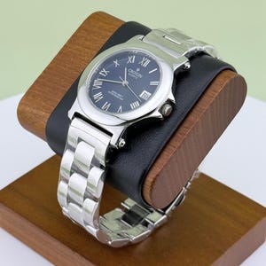 May include: A Croton Quartz wristwatch with a silver band and a dark blue face. The watch features Roman numeral hour markers and a date display. The watch is displayed on a wooden stand with a black leather cushion.