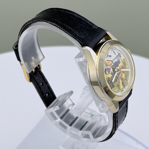 K&ouml;nnte beinhalten: Eine Vintage-Armbanduhr mit goldfarbenem Geh&auml;use und schwarzem Lederarmband. Das Zifferblatt der Uhr zeigt eine farbenfrohe Illustration. Die Uhr wird auf einem transparenten Kunststoffst&auml;nder ausgestellt.