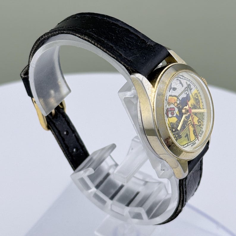 K&ouml;nnte beinhalten: Eine Vintage-Armbanduhr mit goldfarbenem Geh&auml;use und schwarzem Lederarmband. Das Zifferblatt der Uhr zeigt eine farbenfrohe Illustration. Die Uhr wird auf einem transparenten Kunststoffst&auml;nder ausgestellt.
