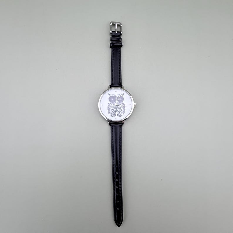 May include: A black leather strap watch with a silver-colored face. The watch face features a detailed owl design. The watch has a silver-colored case and buckle.