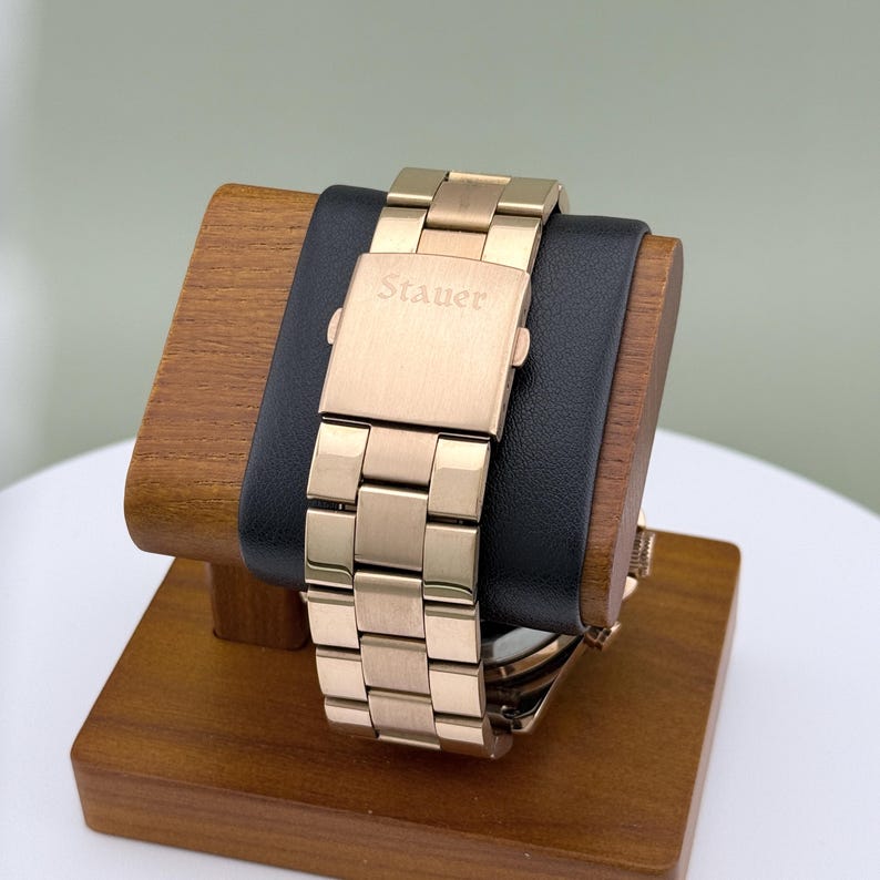 K&ouml;nnte beinhalten: Eine goldfarbene Stauer-Uhr mit rechteckigem Verschluss, pr&auml;sentiert auf einem Holzst&auml;nder mit einem schwarzen Lederkissen. Das Uhrenarmband besteht aus rechteckigen Gliedern. Das Wort "Stauer" ist auf dem Verschluss eingraviert.
