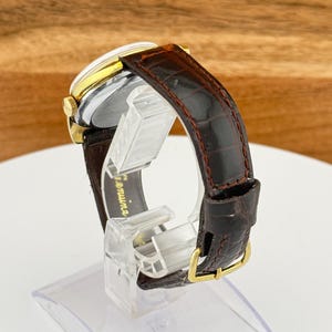 May include: A vintage wristwatch with a gold-toned case and a brown leather band. The watch has a silver face and a dark brown, textured leather strap with a gold-colored buckle. The watch is displayed on a clear stand.