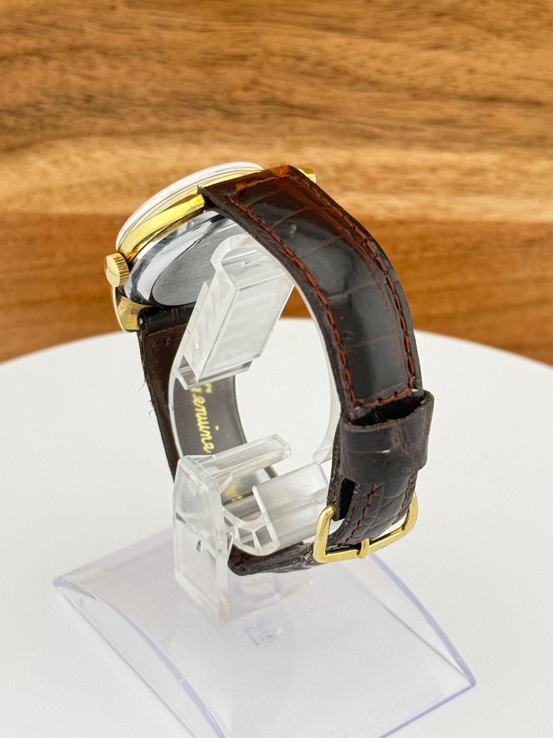 May include: A vintage wristwatch with a gold-toned case and a brown leather band. The watch has a silver face and a dark brown, textured leather strap with a gold-colored buckle. The watch is displayed on a clear stand.