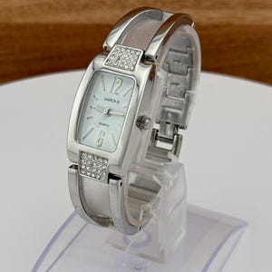 May include: Silver-toned watch with a rectangular face and a band-style bracelet. The watch face is light-colored with the text "watch-it" and the numbers 12 and 6. The band features decorative rhinestones.