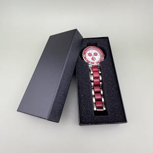 May include: A red and silver wristwatch with a white face and silver hands is displayed in an open black box. The watch has a red bezel and a red band. The box is rectangular and has a textured surface.