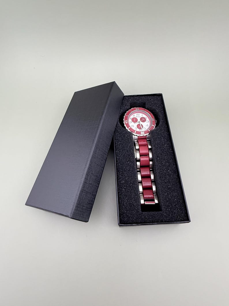 May include: A red and silver wristwatch with a white face and silver hands is displayed in an open black box. The watch has a red bezel and a red band. The box is rectangular and has a textured surface.