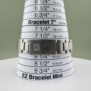 Puede incluir: Un brazalete de reloj de metal plateado se muestra en una herramienta de medici&oacute;n blanca EZ Bracelet Mini. El brazalete mide 17,8 cm en la herramienta. El cierre presenta un s&iacute;mbolo de cruz.
