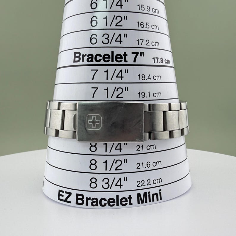 Puede incluir: Un brazalete de reloj de metal plateado se muestra en una herramienta de medici&oacute;n blanca EZ Bracelet Mini. El brazalete mide 17,8 cm en la herramienta. El cierre presenta un s&iacute;mbolo de cruz.
