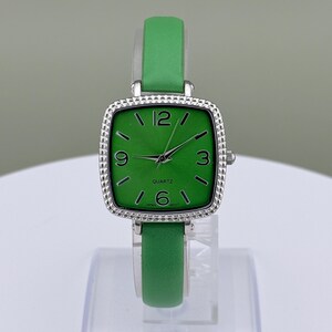 May include: A green quartz watch with a square face and a silver bezel. The watch has a green leather strap and displays the numbers 12, 3, 6, and 9. The watch face reads "QUARTZ" and "JAPAN MOV'T".