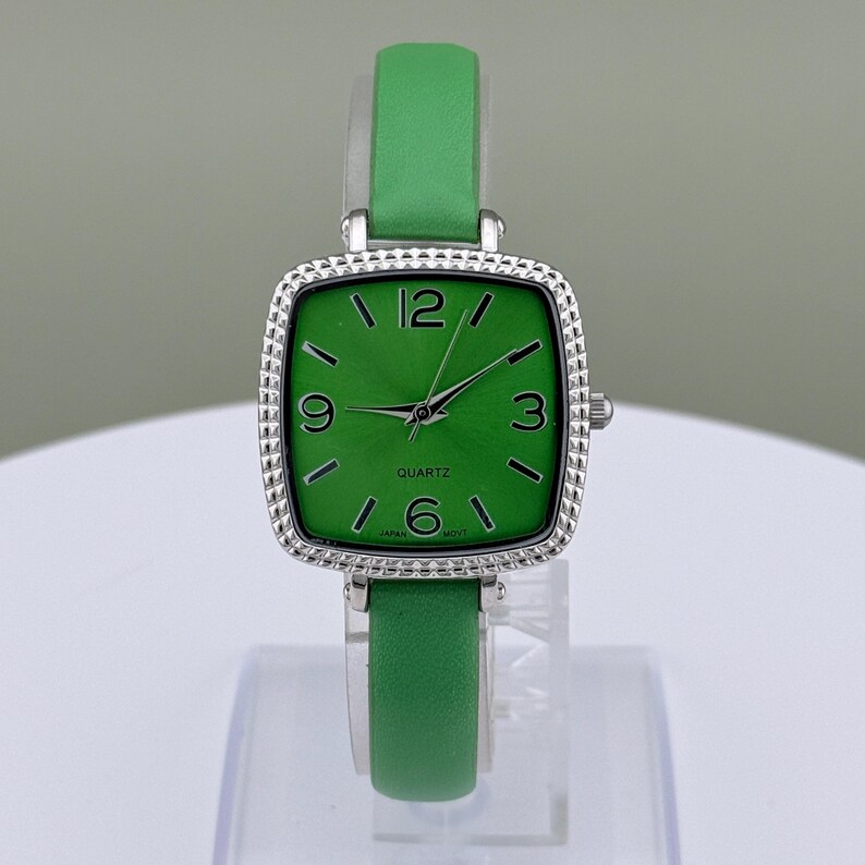May include: A green quartz watch with a square face and a silver bezel. The watch has a green leather strap and displays the numbers 12, 3, 6, and 9. The watch face reads "QUARTZ" and "JAPAN MOV'T".