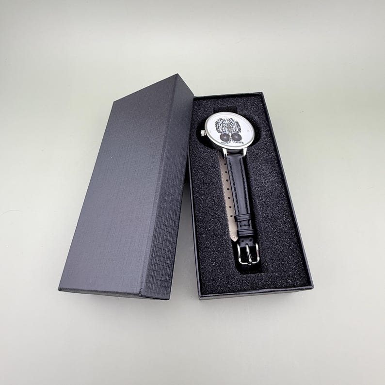 May include: A black leather strap watch with a silver-colored case and a white face featuring a detailed design. The watch is presented in an open black gift box with a textured exterior and a foam interior.
