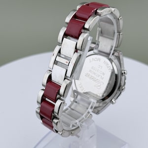 May include: A silver-toned wristwatch with a burgundy band. The watch features a rectangular face and a stainless steel band with burgundy links. The watch is displayed on a clear stand.